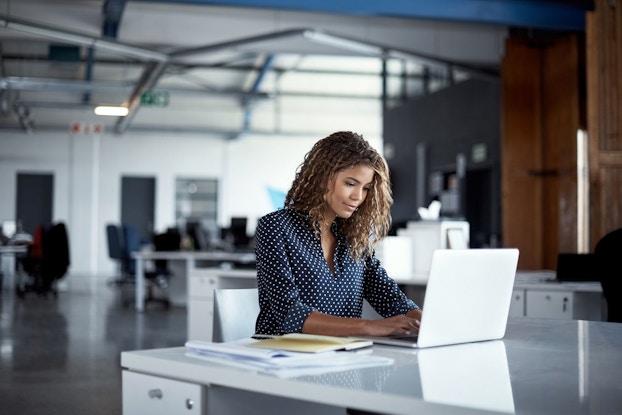 A woman employee sits at a desk and types on a laptop computer. Behind her is a modern, open-plan office space.