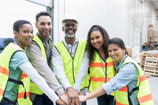  A group of employees of various ages, races, and genders stands in a semicircle with their hands together in the middle. They wear neon vests and smile for the camera.