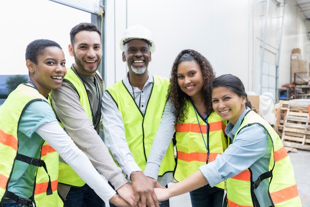 A group of employees of various ages, races, and genders stands in a semicircle with their hands together in the middle. They wear neon vests and smile for the camera.