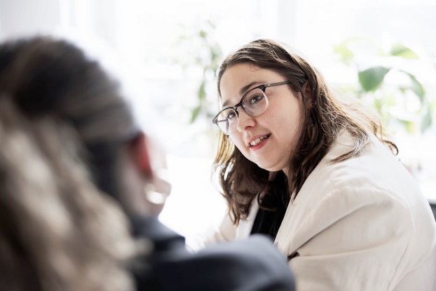  Two women are pictured in an office setting. One young professional woman smiles as she listens to her coworker.