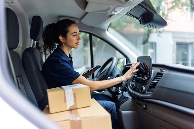  Employee in her car typing into the navigation system with packages to deliver on the passenger seat.