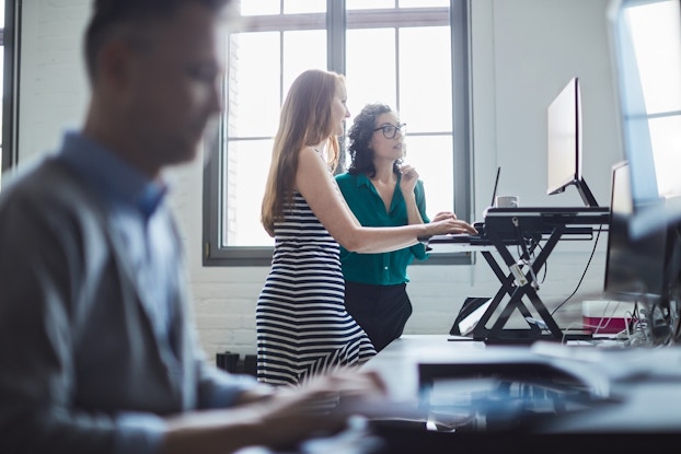 Two woman stand in front of a laptop on top of a stand that converts their workspace into a standing desk. In the foreground, a man, out of focus, sits in front of his own laptop.