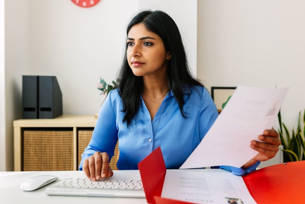 A woman human resources manager is seated at a desk and looks at her computer screen while holding a job candidate's resume.
