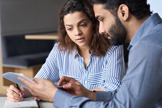 Two employees, a male and a female, sitting next to each other review some paperwork. The man points to one of the papers he is holding.