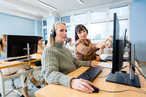 A woman manager trains a new employee at a call center. The manager points to the computer screen to illustrate a point for the female trainee.