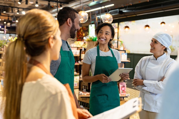 Four employees stand in a loose circle in the middle of a store. The two employees in the middle, a man and a woman wearing green aprons, are looking at each other and smiling. The woman is holding an electronic tablet. The other two employees, a woman in a chef's uniform, and a woman facing away from the viewer, are also looking at the two employees in the middle. The store in the background is a large space with freestanding shelves holding boxed and bottled goods.