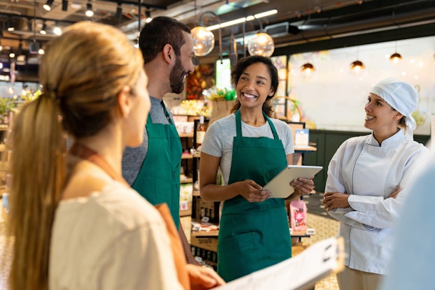  Four employees stand in a loose circle in the middle of a store. The two employees in the middle, a man and a woman wearing green aprons, are looking at each other and smiling. The woman is holding an electronic tablet. The other two employees, a woman in a chef's uniform, and a woman facing away from the viewer, are also looking at the two employees in the middle. The store in the background is a large space with freestanding shelves holding boxed and bottled goods.