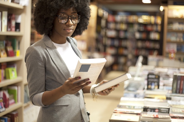  A young woman stands in a bookstore and considers two books, one in each hand. The woman is dressed business casual, in a white blouse and gray blazer.
