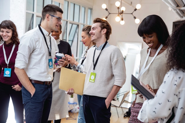 A group of young businesspeople mingle and converse happily in a large minimalist room. The people are wearing business casual clothing and lanyards with blue or light green ID cards hanging from them.