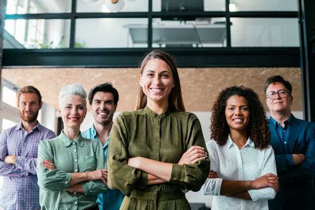 Six people of various ages, genders and races stand in a group with their arms folded. They're all wearing button-up shirts in a variety of colors and they have smiles on their faces.