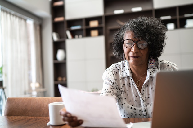 A middle-aged woman wearing glasses and a black-and-white blouse sits at a wooden table and considers the piece of paper that she holds in her hand. In the background is a living room or den, with dark wood shelving along the back wall. An open laptop sits in the foreground, in front of the woman.