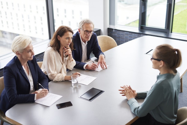 A young woman with her hair in a high ponytail sits across a conference room table from two older women and an older man, all wearing business casual clothing.