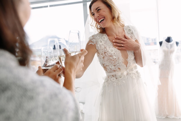  A laughing woman in a wedding dress clinks glasses of champagne with friends off-camera. In the background is a wall of windows and another dress on a mannequin.