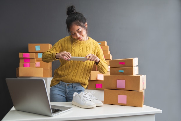  A woman in a bright yellow sweater takes a picture of a pair of white sneakers with her smartphone. She is surrounded by labelled cardboard boxes and an open laptop.