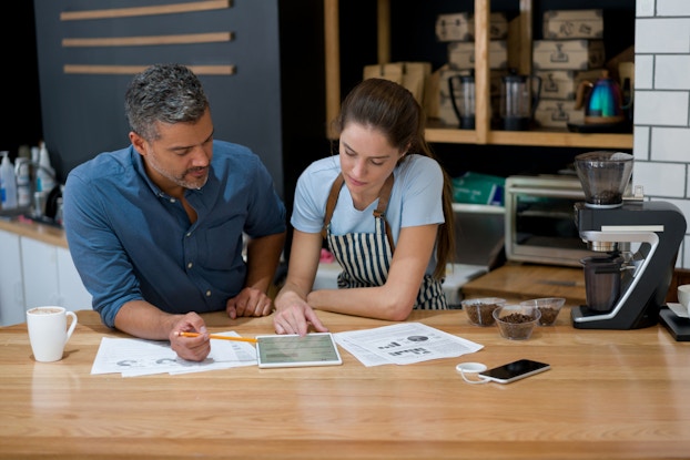 A middle-aged man and a younger woman lean on a countertop and look at the screen of an electronic tablet. Various papers on the counter show bar and pie charts. Behind and next to the man and woman are cups of coffee beans, a coffee machine, and shelves holding wooden boxes and coffee carafes.