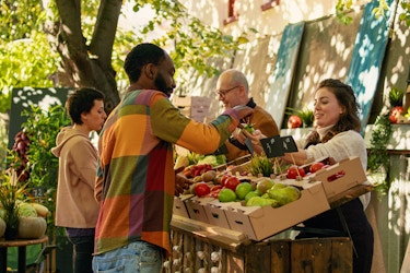 Two customers stand in front of an angled table holding boxes of fruit and vegetables. The produce stand is outside in the shade of a tree. Each customer is interacting with an employee standing behind the table. The buyer and seller closest to the camera are a man in a multi-colored checkered shirt and a woman in a cream-colored sweater, respectively. The man takes a small pear out of the woman's hand. The other buyer and seller in the background are a short-haired woman in a tan hoodie and an older man with glasses. The produce on the table includes potatoes, apples, pears, and tomatoes.