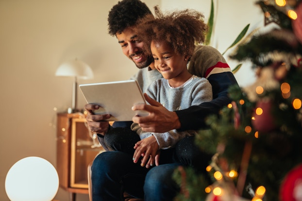  Father and child smiling inside their home decorated for the holidays looking at a tablet.