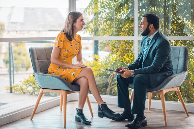 A woman and a man sit in chairs facing each other, talking. The woman wears a yellow dress and black ankle boots. She has her legs crossed and is leaning slightly forward. The man is wearing a dark blue suit with a green-and-white-striped tie. He is holding an electronic tablet. Behind the chairs, two banks of windows meet to form the corner of a room. Outside the floor-to-ceiling windows is a garden filled with trees and shubbery.