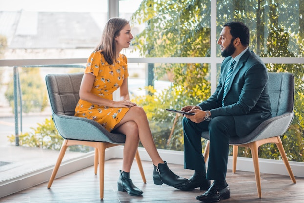  A woman and a man sit in chairs facing each other, talking. The woman wears a yellow dress and black ankle boots. She has her legs crossed and is leaning slightly forward. The man is wearing a dark blue suit with a green-and-white-striped tie. He is holding an electronic tablet. Behind the chairs, two banks of windows meet to form the corner of a room. Outside the floor-to-ceiling windows is a garden filled with trees and shubbery.