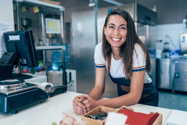 A young woman leans over the counter of her eating establishment and smiles at the viewer.