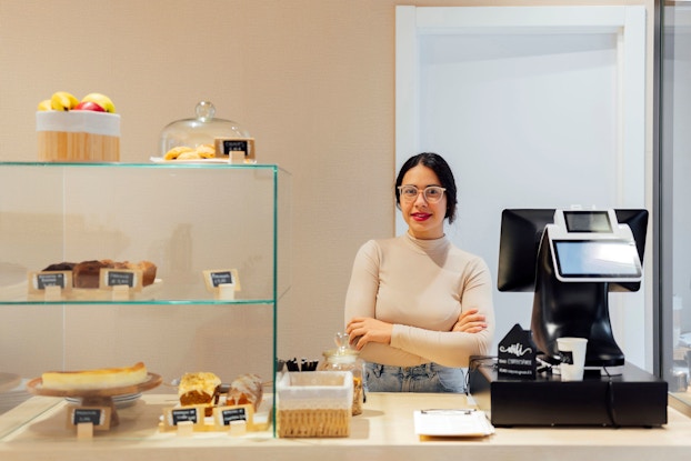A young woman bakery owner stands behind the point-of-sale kiosk at her shop. She is smiling at the viewer.