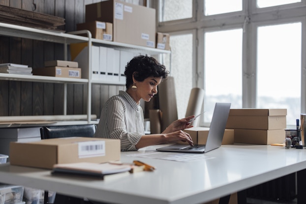  Person working inside an office at a desk with shelves of boxes behind her.