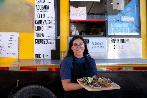  A woman in glasses and a black apron smiles proudly while holding a wooden platter of gourmet tacos garnished with lime slices. Behind her is a large yellow food truck. Attached to the side of the truck are several menus with the names of Mexican foods, such as pollo, tripa and chorizo, written in bold black type, and descriptions of the food written in smaller text. A smaller sign reads "Ya abrimos! We are open again!"
