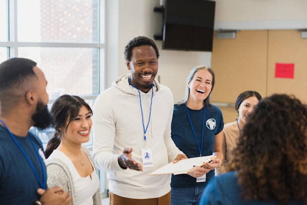 Five casually dressed people wearing blue lanyards stand in a line smiling. The lanyards hold white cards bearing a blue symbol and the word "VOLUNTEER." In the foreground, a sixth person, who has long, curly, dark hair, stands facing away from the viewer and toward the group of people.
