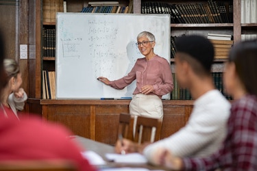  A woman with short white hair and glasses stands and lectures in front of a whiteboard covered with equations written in marker. Out of focus in the foreground are several young people sitting in chairs around a table. In the background are wooden shelves of folders and books. 