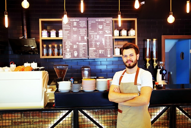  A man wearing an apron stands in front of a coffee shop counter, behind which is mounted a menu.