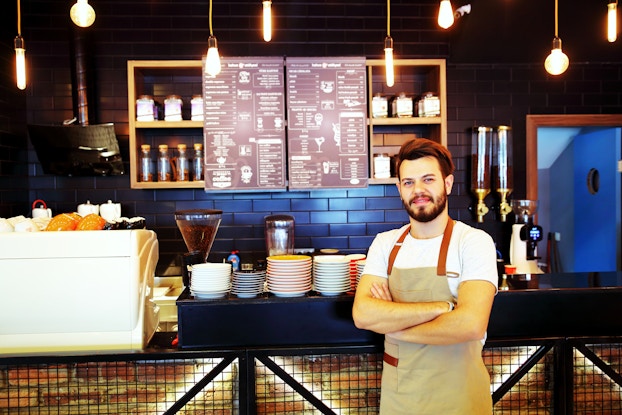 A man wearing an apron stands in front of a coffee shop counter, behind which is mounted a menu.
