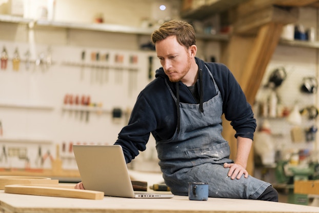  A man in a woodworking workshop sits on the edge of a wooden table and looks down at an open laptop. The man is wearing a stained blue apron over a dark blue hoodie. In the background, out of focus, are racks and shelves holding tools for carving and cutting.