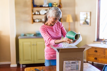  A smiling older woman stands in her home and removes bubble wrap that was wrapped around a green ceramic pitcher. On the table in front of the woman is an open cardboard box with a mailing label on the front. The woman has gray hair pulled back into a ponytail, and she wears a pink shirt with white stripes. 