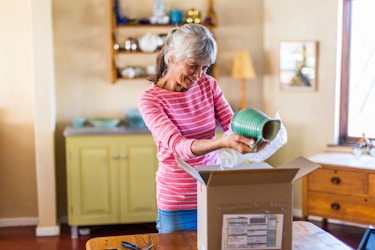 A smiling older woman stands in her home and removes bubble wrap that was wrapped around a green ceramic pitcher. On the table in front of the woman is an open cardboard box with a mailing label on the front. The woman has gray hair pulled back into a ponytail, and she wears a pink shirt with white stripes.