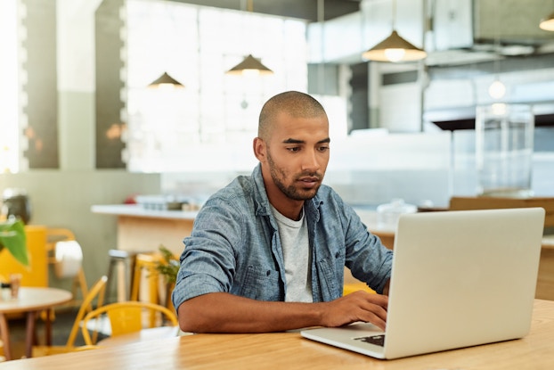  A young man sits at a table in an empty but brightly lit cafe and types on a laptop. The man has a shaved head and a dark beard, and he wears a chambray shirt over a white T-shirt.