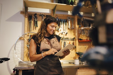  A woman stands in a workshop, leaning against a chrome-topped counter and looking down at the electronic tablet in her hands. She has long brown hair and wears a black T-shirt under a brown leather work apron. Behind her is a wall covered with mounted and hanging tools, including wrenches and hammers. The silver spokes of a bike tire sits on top of the counter. 