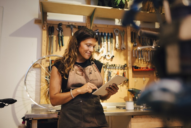  A woman stands in a workshop, leaning against a chrome-topped counter and looking down at the electronic tablet in her hands. She has long brown hair and wears a black T-shirt under a brown leather work apron. Behind her is a wall covered with mounted and hanging tools, including wrenches and hammers. The silver spokes of a bike tire sits on top of the counter.