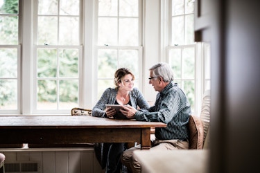  Father and adult daughter sitting at a kitchen table looking at a tablet. 