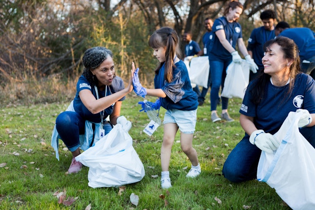  People in dark blue shirts pick up trash in a grassy field. In the background, against a forest backdrop, participants hold open white trash bags. In the foreground, a little girl high-fives a woman holding a trash bag. Another woman, at right, watches the high-five and swallows.