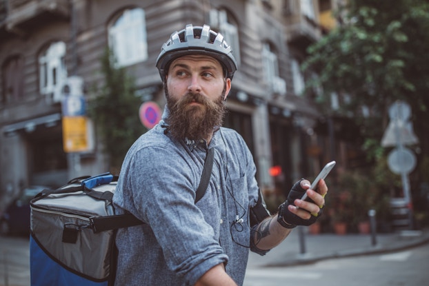  A bearded man in a city street intersection looks off-camera. He is carrying a cellphone and wearing a helmet and a large square backpack for carrying food. The photo is shot from the waist up, but the man is presumably sitting on a bike.