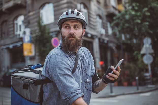 A bearded man in a city street intersection looks off-camera. He is carrying a cellphone and wearing a helmet and a large square backpack for carrying food. The photo is shot from the waist up, but the man is presumably sitting on a bike.