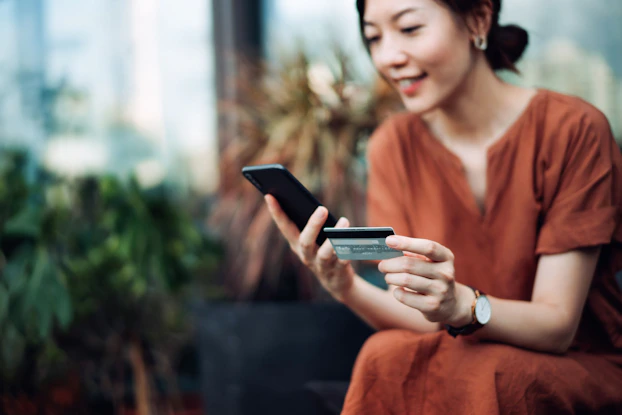 A young Asian woman is sitting outside on a patio. She is holding a smartphone in her left hand a credit card in her right hand. She is preparing to make an online payment using her credit card.