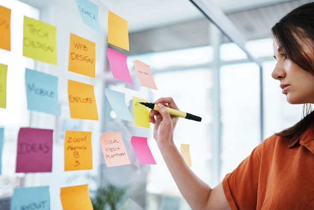  A woman in an orange blouse uses a fine-point marker to write on a yellow post-it note stuck on a glass partition. The yellow note is placed in a grid of other post-it notes of varying colors. Words and phrases such as "Ideas," "Explore" and "Web Design" are written on the notes.