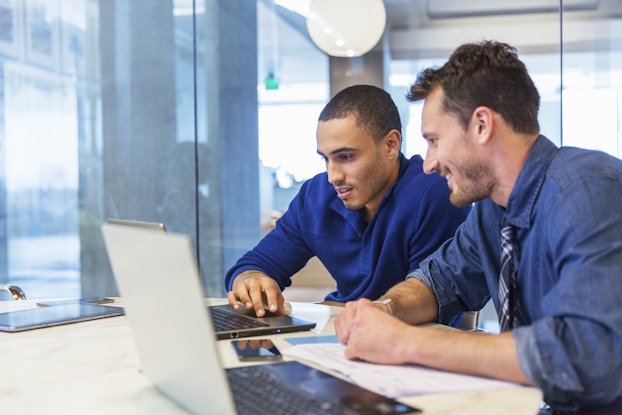 Two men sit side by side at a desk looking at the screen of an open laptop. The man on the left is wearing a midnight blue button-up shirt and has a relaxed look on his face. His right hand is operating the laptop trackpad. The man on the right is wearing a blue button-up shirt and gray plaid tie and has a smile on his face. Another open laptop sits on the table in the foreground, out of focus.