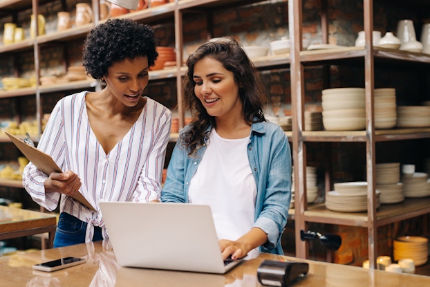 Two women stand at a table, looking at the screen of an open laptop. The woman on the left has a small afro and wears a gray-and-white-striped clouse. She holds a clipboard. The woman on the right has medium-length brown hair and wears a chambray shirt over a white T-shirt. She is typing on the laptop. Both women are smiling with their mouths slightly open, as if they are speaking or laughing.The room behind them is a medium-sized warehouse room with a large shelving unit filled with ceramic bowls and cups.