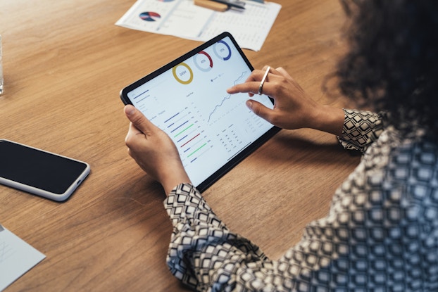 An over-the-shoulder shot of a woman examining charts and data on the screen of a tablet.