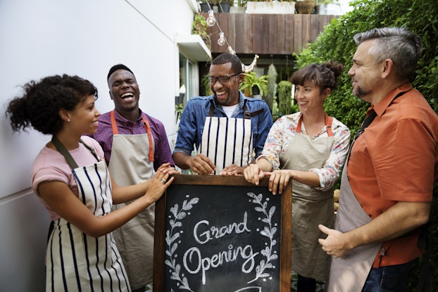  A group of five people wearing aprons stand around a chalkboard sign. The sign reads "Grand Opening" and the words are surrounded by a laurel wreath drawn in chalk. The five people are of different races and genders, and they stand with their hands on top of the sign, facing each other and smiling or laughing in celebration.