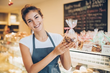A woman bakery store owner smiles at the viewer. She is holding up a exquisitely wrapped package of baked goods.