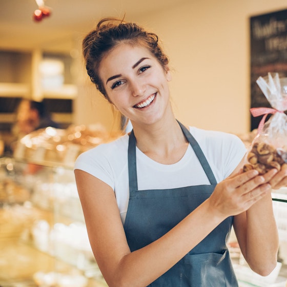 A woman bakery store owner smiles at the viewer. She is holding up a exquisitely wrapped package of baked goods.