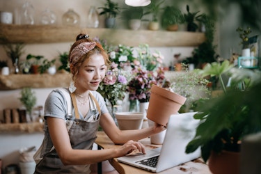  A young woman sits at a wood-topped counter in a plant shop and looks at something on the screen of an open laptop. With one hand, the woman holds a clay pot; with the other, she manipulates the track pad on the laptop. The woman wears a gray apron over a light blue T-shirt. Her hair is pulled up in a bun and partially covered by a paisley-patterned headband. Most of the counter is covered with potted plants and flowers, as are the wooden shelves on the wall behind the woman. 